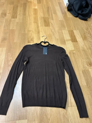 Brun stickad tröja från Lager 157 - Mörkbrun stickad tröja från Lager 157 med rund halsringning och långa ärmar. Tröjan har en enkel, stilren design och ribbade muddar vid ärmslut och nederkant. Oanvänd och helt ny med prislapp.