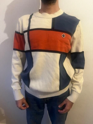 Färgglad stickad tröja med blockmönster - Säljer en stickad tröja med grafiskt blockmönster i vitt, marinblått och rött. Tröjan har rund halsringning, långa ärmar och ribbade muddar. Liten broderad logga på bröstet. Perfekt för dig som gillar retro och streetwear vibes.