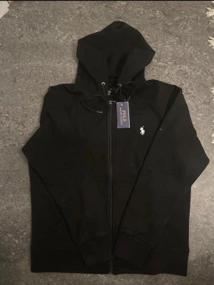 Svart hoodie från Polo Ralph Lauren - Svart hoodie med dragkedja från Polo Ralph Lauren. Klassisk design med vit broderad logga på bröstet, justerbar huva med snören och fickor framtill. Tillverkad i mjukt bomullsmaterial som känns skönt mot huden. Perfekt för en avslappnad och stilren look.