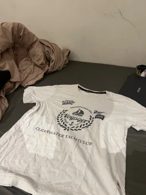 Vit t-shirt Voyager Clearwater Yachtclub M-L - Vit t-shirt från Voyager med tryck framtill: 'Clearwater Yachtclub', segelbåt och amerikansk flagga. Klassisk passform med rund hals och korta ärmar. Perfekt för en avslappnad och stilren look.