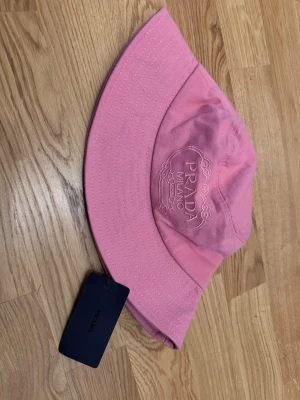 Rosa bucket hat från Prada - Snygg rosa bucket hat från Prada med broderad logga framtill och klassisk bred skärm. Hatten är tillverkad i bomull och har vita sömmar som kontrast. Märkesetikett och originaltagg sitter kvar. Perfekt accessoar för en trendig look.