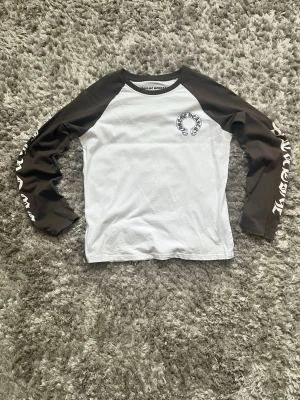 Chrome Hearts långärmad topp - Säljer en långärmad topp från Chrome Hearts med vit kropp och mörkbruna ärmar. Tryck med Chrome Hearts-logga på bröstet och stort korsmotiv på ryggen. Ärmarna har cool textprint längs sidorna. Klassisk baseballmodell i mjuk bomull.