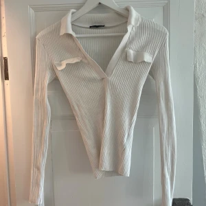Vit ribbad långärmad polotopp - Trendig vit ribbad topp med lång ärm och djup v-ringning. Polokrage och dekorativa fickdetaljer framtill ger en cool touch. Perfekt att styla med jeans eller kjol för en clean look. Perfekt basic topp!