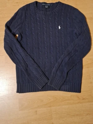 Mörkblå kabelstickad tröja Ralph Lauren - Kabelstickad mörkblå tröja från Ralph Lauren i 100% bomull. Klassisk rund halsringning, långa ärmar och ribbade muddar. Ikoniska vita Polo-loggan broderad på bröstet. Perfekt för dig som gillar stilrena och tidlösa plagg.
