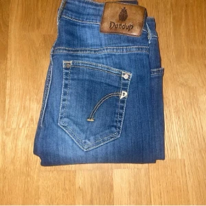Blå jeans från Dondup med raka ben - Säljer ett par klassiska blå jeans från Dondup med raka ben och snygga kontrastsömmar. Jeansen har fem fickor, normal passform och ett brunt läderpatch med logga bak i midjan. Perfekta till din vardagsstil!