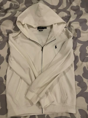 Vit hoodie från Polo Ralph Lauren - Snygg vit hoodie från Polo Ralph Lauren med dragkedja framtill och klassisk logga broderad på bröstet. Hoodien har huva, fickor på sidorna och ribbade muddar vid ärmar och nederkant. Finns inga defekter o sitter bra. De är storlek M men passar perfekt som S. Kom med frågor o prisförslag