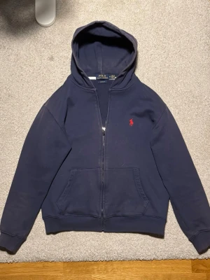 Mörkblå hoodie från Polo Ralph Lauren - Klassisk Ralph Lauren zip-tröja i marinblå färg. Mjuk och skön, med diskret logga på bröstet. Bra skick, inga fläckar eller skador. Jag har tyvärr tappat bort banden till luvan, men kan skicka med dem om jag hittar dem. Vill egentligen inte sälja den, men släpper den för bra pris 🤝  (Passar mer S än M)