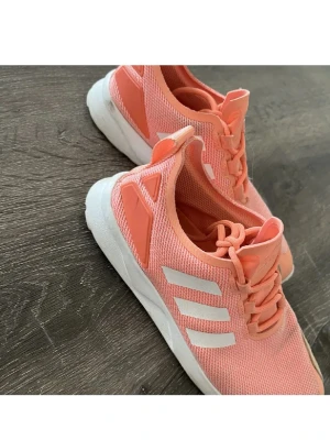 Adidas sneakers i korall och vitt - Snygga Adidas sneakers i korallrosa med vita detaljer och klassiska tre ränder på sidan. Ovandelen är i mesh för bra andningsförmåga och skon har snörning samt vit platt sula. Perfekta för en sportig och trendig look. Fint skick.