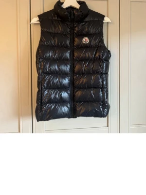 Svart dunväst från Moncler - Snygg svart dunväst från Moncler med hög krage och dragkedja framtill. Västen har ett glansigt material och det klassiska Moncler-logotypen på bröstet. Jag har köpt den här på Plick och den förra ägaren köpte den i en moncler butik i Paris. 