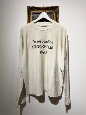 Vit långärmad t-shirt Acne Studios - Vit långärmad t-shirt från Acne Studios med svart tryck på bröstet där det står 'Acne Studios STOCKHOLM 1996'. Tillverkad i mjuk bomull med avslappnad passform och rund hals. Perfekt för en clean och stilren look.