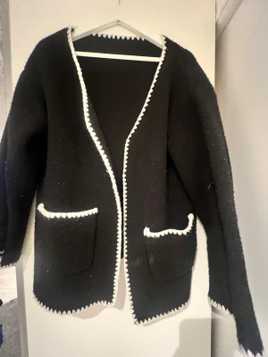 Svart kofta med vita detaljer - Säljer en svart kofta med vita, sicksack-formade kanter längs öppningen, fickorna och nederkanten. Koftan har två stora fickor framtill och är långärmad. Perfekt för dig som gillar en stilren men ändå unik look.