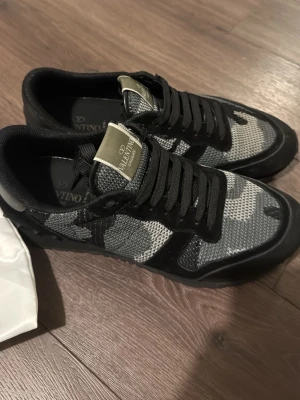 Valentino Rockrunner sneakers svart/grå - Säljer ett par Valentino Rockrunner sneakers i svart och grått med camouflagemönster. Skorna har snörning, detaljer i mocka och textil samt en robust gummisula. Ikonisk logga på plösen och cool streetwear-vibe. Perfekta för dig som vill sticka ut med exklusiva sneakers.