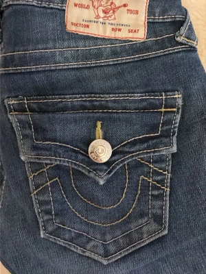 True Religion Joey Low Rise Flare Jeans - Säljer ett par blå True Religion Joey jeans med låg midja och flare-ben. Klassiska kontrastsömmar, snygga fickdetaljer och silverknappar. Tillverkade i slitstarkt jeansmaterial