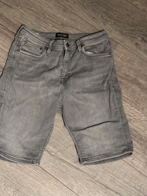 Grå slim fit jeansshorts Jack & Jones - Snygga grå jeansshorts från Jack & Jones i slim fit-modell. Klassisk femficksdesign med bälteshällor och knappgylf. Perfekta för dig som gillar en stilren och enkel look till sommaren.