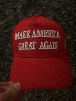 Röd keps med Make America Great Again - Röd keps med vit brodyrtext 'MAKE AMERICA GREAT AGAIN' framtill och broderad amerikansk flagga på sidan. Klassisk böjd skärm och justerbar passform. Perfekt statement-accessoar för dig som vill sticka ut.