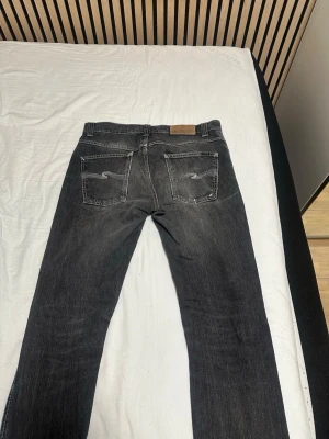 Svarta Nudie Jeans Co - Säljer ett par svarta nudie jeans co med klassiska fyra fickor och snygga kontrastsömmar. Jeansen har en mörk tvätt och N-logga på bakfickan. Perfekta för dig som gillar en avslappnad men ändå stilren look.