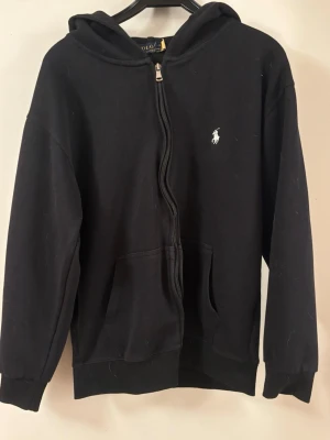 Svart hoodie från Polo Ralph Lauren - Klassisk svart hoodie från Polo Ralph Lauren med dragkedja framtill och vit broderad logga på bröstet. Hoodien har huva, känguruficka och ribbade muddar vid ärmslut och nederkant. Tillverkad i mjuk bomullsblandning för skön komfort. Säljer pga för liten storlek