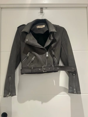Grå croppad skinnjacka med bälte - Snygg grå croppad skinnjacka från Zara med flera dragkedjor, bred krage och bälte i midjan. Jackan har långa ärmar med dragkedjor vid ärmsluten och coola detaljer som axelklaffar. Perfekt för en edgy streetstyle-look.