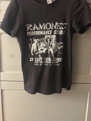 Svart Ramones t-shirt XS - Säljer en svart t-shirt från Ramones i storlek XS. T-shirten har ett stort vitt tryck framtill med texten 'Ramones Performance Studio' och en bild på bandet. Klassisk rund hals och korta ärmar. Perfekt för dig som gillar bandtröjor och vill ha en cool look.