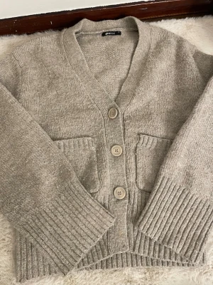 Beige stickad kofta från Gina Tricot - Mysig beige stickad kofta från Gina Tricot i storlek XS. Koftan har tre stora knappar framtill, två fickor och breda ribbade muddar vid ärmslut och nederkant. Perfekt för lager på lager under kyliga dagar.