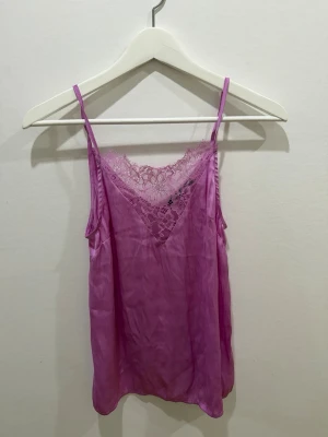 Rosa linnetopp med spets från Gina Tricot - Säljer en rosa linnetopp från Gina Tricot med tunna axelband och v-ringning dekorerad med spets. Toppen har en lätt glansig finish och är perfekt för att skapa en trendig lager-på-lager-look.