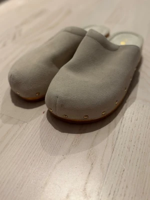 Beige mocka träskor från LUX - Snygga beige sandaler från LUX i mjuk mocka med träbotten och guldfärgade nitar längs sidorna. Skorna har rund tå och öppen häl, perfekt för dig som gillar klassisk träskodesign med modern touch.