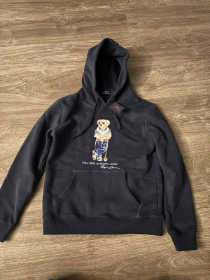 Svart hoodie Polo Bear Ralph Lauren - Snygg svart hoodie från Ralph Lauren med Polo Bear-tryck på bröstet. Klassisk känguruficka och dragsko i huvan. Mjuk bomullskvalitet och avslappnad passform, perfekt för en chill och stilren look.