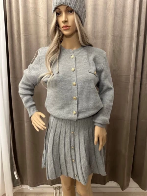Grått stickat set med kjol och topp - Snyggt grått stickat set med långärmad topp och matchande plisserad kjol. Toppen har guldknappar och dekorativa fickor. Setet är mjukt och bekvämt, perfekt för en trendig look.