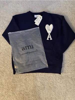Mörkblå stickad tröja från Ami - Mörkblå stickad tröja från Ami Alexandre Mattiussi med vit hjärtlogga och bokstaven A på bröstet. Tröjan har rund halsringning och ribbade muddar vid ärmslut och nederkant. Perfekt för dig som gillar stilrena detaljer och trendiga plagg.