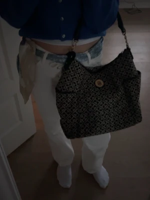 Mönstrad axelväska från Tommy Hilfiger - Säljer en snygg axelväska från Tommy Hilfiger med svart och beige geometriskt mönster och guldfärgad TH-logga framtill. Väskan har en mjuk form och justerbar axelrem. Perfekt för dig som vill ha en trendig och rymlig väska till vardagen. Får plats med MacBook och näst intill oanvänd!