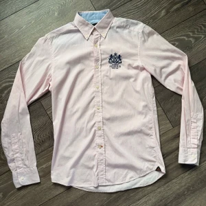 Rosa och vit randig skjorta Morris - Snygg rosa och vit randig skjorta från Morris med klassisk krage och broderad logga på bröstet. Skjortan har långa ärmar och knäppning framtill. Perfekt för dig som gillar stilrena detaljer och tidlös design.