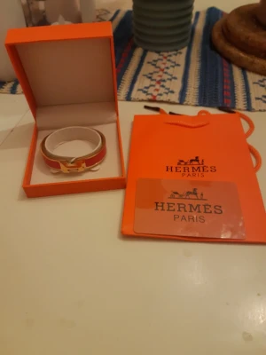 Rött armband från Hermès - Snyggt rött armband från Hermès med guldfärgad metall och klassisk H-detalj framtill. Armbandet har en elegant och stilren design med vit insida och kommer i originalask och påse. Perfekt accessoar för att lyfta din outfit.