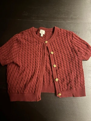 Vinröd kabelstickad kofta H&M - Kortärmad vinröd kabelstickad kofta från H&M med rund halsringning och dekorativa guldfärgade knappar framtill. Ribbstickade muddar vid ärmslut och nederkant ger en snygg finish. Perfekt att styla över en topp eller klänning.