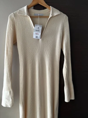 Ribbad långklänning från Bershka - Supersnygg ribbad långklänning från Bershka i en ljus beige ton. Klänningen har v-ringning, krage och långa ärmar. Materialet är stretchigt och mjukt, perfekt för en trendig look. Enkel men ändå stilren design som är lätt att matcha.