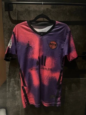PSG matchtröja lila/rosa Jordan - Säljer en Paris Saint-Germain matchtröja i lila och rosa med svart logga och detaljer. Tröjan har korta ärmar, V-ringning och PSG-märke på bröstet samt Ligue 1-märke på ärmen. Tillverkad i lätt och ventilerande material, perfekt för fotboll eller träning.