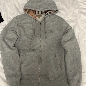 Grå zip hoodie från Burberry med dragkedja - Grå hoodie från Burberry i bomull med klassiskt rutigt foder i huvan. Tröjan har dragkedja framtill, snörning vid huvan och två stora fickor. Ikonisk Burberry-logga broderad på bröstet. Perfekt för en chill och stilren look.