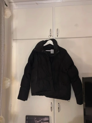 Svart pufferjacka från Etirel - Snygg svart pufferjacka från Etirel med hög krage men tyvärr ingen dragkedja utan bara knappar att stänga men funkar lika bra. Jackan har två stora fickor, kom till användning förut men inte längre.