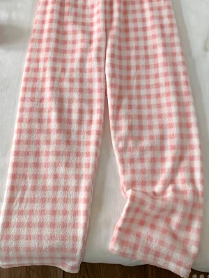 Rosa rutiga fleecebyxor loungewear - Supermysiga loungewear-byxor i fluffig fleece med rosa och vitt rutmönster. Byxorna har resår i midjan och raka ben, perfekta för chill hemma. Materialet är mjukt och värmande, perfekt för lata dagar eller kvällar i soffan.