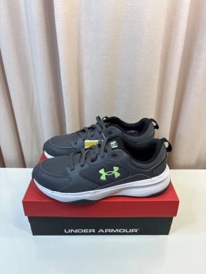 Under Armour - Snygga grå sneakers från Under Armour med vit sula och limegrön logga på sidan. Skorna har snörning och är tillverkade i syntetmaterial med meshpaneler för extra andningsförmåga. Perfekta för dig som gillar sportig stil.