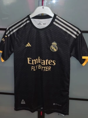Real Madrid svart matchtröja Adidas - Säljer en svart Real Madrid matchtröja från Adidas med gula detaljer och tryck. Tröjan har korta ärmar, klubbmärke på bröstet och VINI JR 7 på ryggen. Perfekt för fotbollsfans som vill stötta sitt lag med stil. Den är använd fåtal gånger. Som ni ser längst ner av tröjan är de lite dåligt. Jag tog ut en tråd. Men man kan lätt sy de så blir den som ny. Inget annat är fel med den och den är ren! Priset är inte hugget i sten. Hör av er om ni har frågor! 