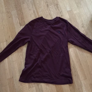 Vinröd långärmad tröja Polo Ralph Lauren - Snygg vinröd långärmad tröja från Polo Ralph Lauren med klassisk rund hals och liten blå logga broderad på bröstet. Tillverkad i mjuk bomull som känns skön mot huden. Perfekt för dig som gillar stilrena och enkla plagg med en touch av premium.