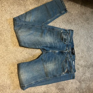 Replay Anbass blå jeans, W30 L32 - Snygga Replay Anbass jeans i klassisk blå tvätt med lätt slitningar och slim passform. Jeansen har fem fickor, dragkedja och tydliga Replay-detaljer på bakfickorna. Perfekt för dig som gillar stilrena och moderna jeans.