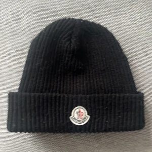Svart ribbstickad mössa från Moncler - Svart ribbstickad mössa från Moncler med klassisk uppvikt kant och broderad logotyp framtill. Mössan är tillverkad i mjuk ull och har en stilren, enkel design som passar perfekt till kalla dagar.