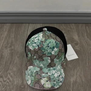 Gucci keps med blommigt mönster - Snygg keps från Gucci med klassiskt GG-mönster och blommor i grönt, vitt och blått. Kepsen har svart baksida och böjd skärm. Perfekt accessoar för att sticka ut med lyxig känsla och trendig design.
