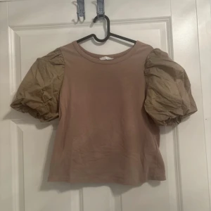 Beige topp med puffärmar från H&M - Säljer en beige topp från H&M med riktigt stora puffärmar i en något ljusare beige nyans. Toppen har rund halsringning och är tillverkad i mjuk bomull med ärmar i ett prassligt material. Perfekt för dig som vill sticka ut med coola detaljer. Använd 1 gång 