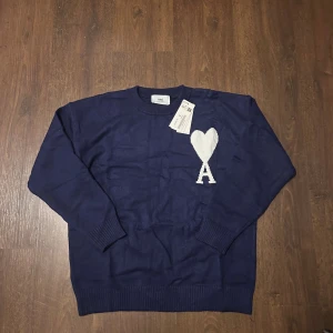 Mörkblå stickad tröja från Ami - Helt ny mörkblå stickad tröja från Ami paris med vit hjärt- och A-detalj på bröstet. Klassisk rund halsringning och ribbade muddar vid ärmslut och nederkant. Perfekt för dig som gillar stilrena och trendiga plagg.