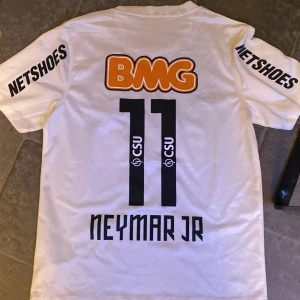 Santos Neymar Jr matchtröja Nike - Säljer en officiell Santos FC fotbollströja från Nike med Neymar Jr och nummer 11 på ryggen. Tröjan är vit med svarta tryck, orange BMG-logga och klubbmärke med två gula stjärnor. Dri-Fit material för skön känsla och bra ventilation. Kortärmad modell med rund hals.