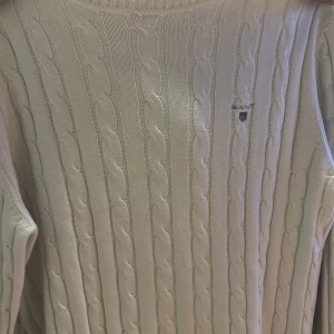 Vit kabelstickad tröja från GANT - Snygg vit kabelstickad tröja från GANT med diskret logga broderad på bröstet. Tröjan har långärmad design och rund hals, perfekt för dig som gillar klassisk och clean stil. Materialet är mjukt och stickat, vilket gör den skön att bära. Skick 10/10 aldrig andvänd. Storlek L men sitter som M. Pris kan diskuteras! Hör av er vid frågor!