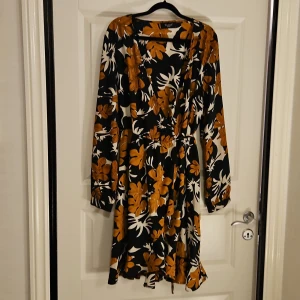 Blommig kortklänning från Sisters Point - Supersnygg kortklänning från Sisters Point med blommigt mönster i svart, vitt och senapsgult. Klänningen har omlott-v-ringning, långa ärmar och knyt i midjan för extra shape. Materialet är mjukt och flowy, perfekt för dig som gillar statement-plagg.