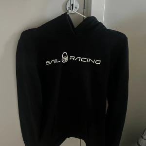 Svart Sail Racing hoodie med tryck - Svart hoodie från Sail Racing med vit logotyptryck på bröstet och en mindre logga på huvan. Tröjan har klassisk huva, lång ärm och känguruficka framtill. Mjuk och bekväm, perfekt för chill dagar. Materialet är bomull och polyester.